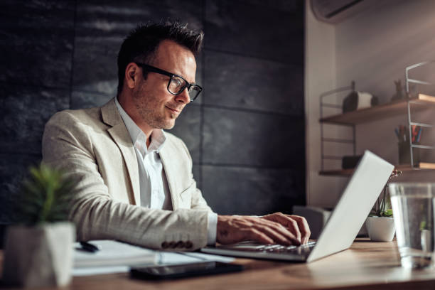 Businessman sitting at his desk and using laptop in the office Businessman wearing linen suit and eyeglasses sitting at his desk by the window and using laptop in the office laptop businessman business men stock pictures, royalty-free photos & images