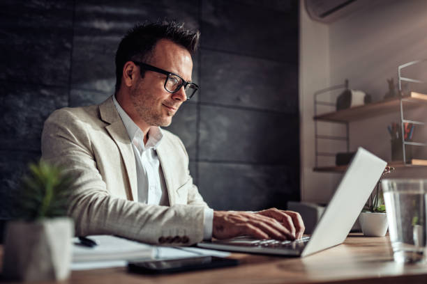businessman sitting at his desk and using laptop in the office - men laptop businessman using laptop imagens e fotografias de stock