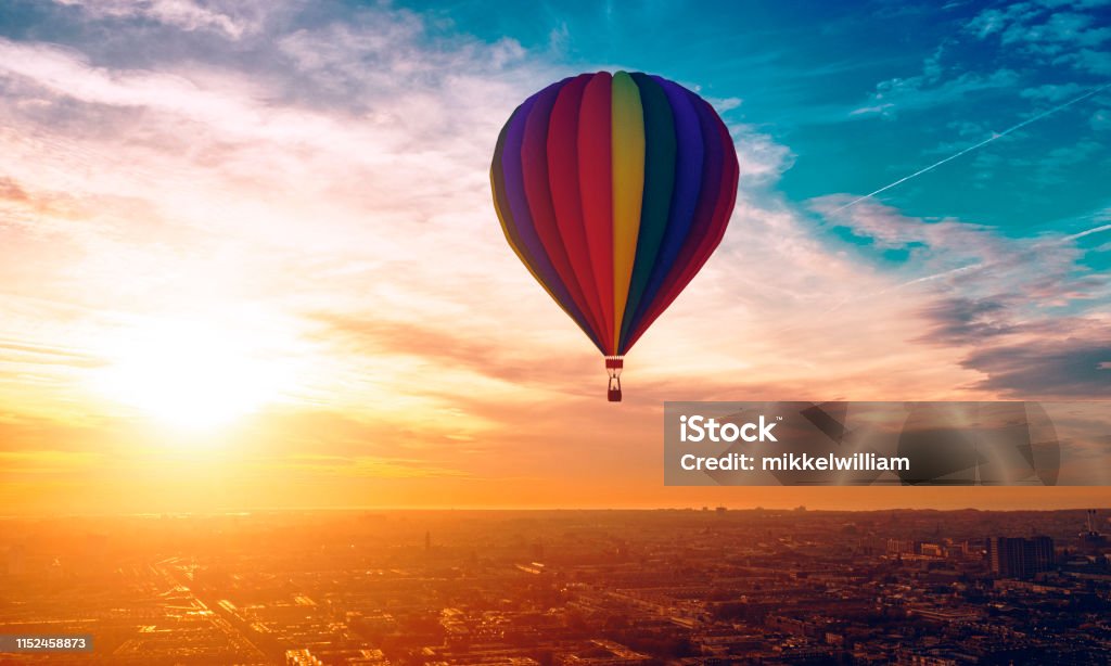 Hot air ballon travels above the city as sun sets in the horizon Hot air ballon with different colors travels across the sky. A city is below and rooftops are visible. 
Note: The hot air ballon is made in a 3D program. Property release attached. Hot Air Balloon Stock Photo