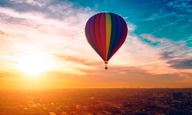 Hot air ballon travels above the city as sun sets in the horizon Hot air ballon with different colors travels across the sky. A city is below and rooftops are visible. 
Note: The hot air ballon is made in a 3D program. Property release attached. hot air balloon photos stock pictures, royalty-free photos & images