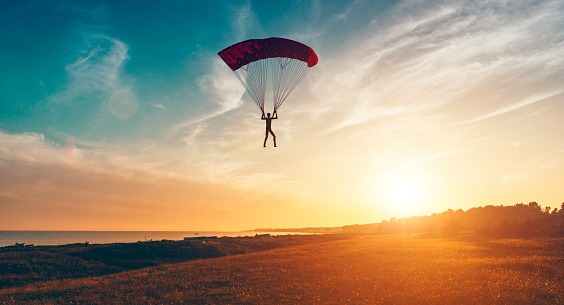 Sun shines and is about to set in the horizon as man parachutes through the air. He is about to land on the ground. 
Note: The man and parachute is made in a 3D program. Property release attached.