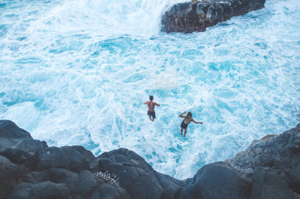 A couple in their 20s leap through the air off a rocky cliff into the white capped ocean waves crashing in far below. The shot is from behind them.