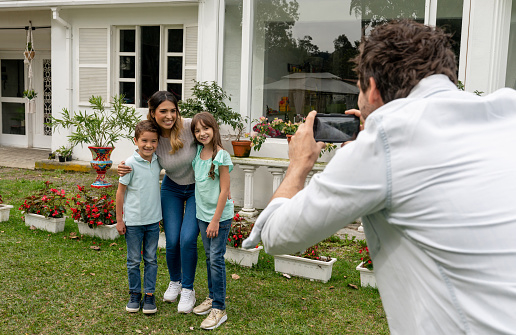 Father taking a picture of their family in front of their new house using a cell phone - lifestyle concepts