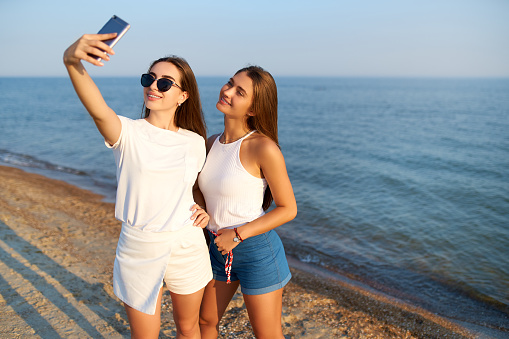 Two young women taking a selfie on the beach with a sea view. Friends are smiling looking at the camera. Girls wearing blue denim. Eco cotton concept.