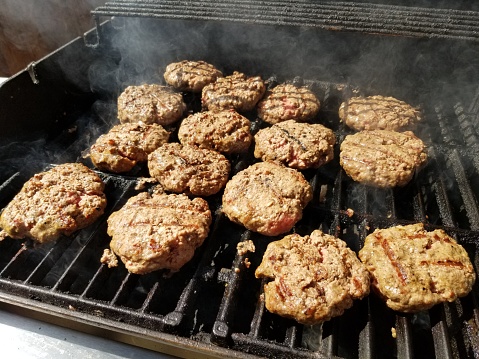 Close-up of several rows of beef burgers with grill marks cooking on an outdoor barbecue grill, May 27, 2019