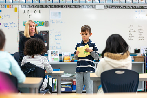 A cute little boy of European descent presents his work to his class. He is reading his assignment while the other students listen attentatively. He looks proud.