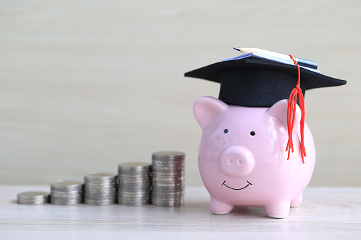 Graduation hat on pink piggy bank with stack of coins money on wooden background, Saving money for education concept
