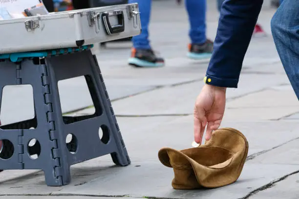 A man putting a pound coin into a busker's leather hat. It has been placed on the paving stones of a busy city centre where shoppers and tourists pass by. The hat is next to a foldaway plastic stool, which has a collection of homemade CD's inside it.