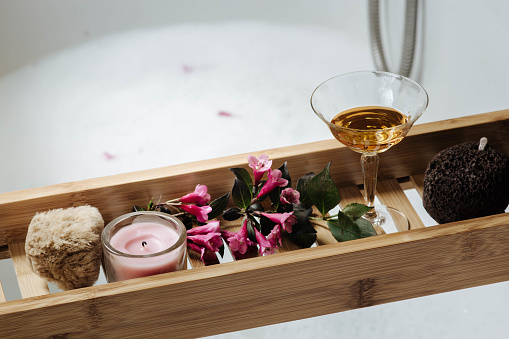 female bathroom - romantic bath with natural pumice, glass of white wine, pink flowers and candle on shelf for a bathtub