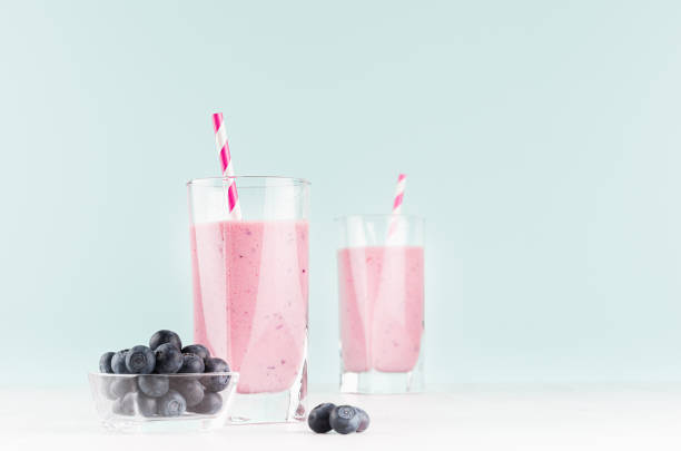 koktajle mleczne jagodowe w przezroczystym szkle z jagodami w misce, słoma w paski na stole z białego drewna i ściana w kolorze miętowym, kopiuj przestrzeń. - blueberry smoothie glass striped zdjęcia i obrazy z banku zdjęć