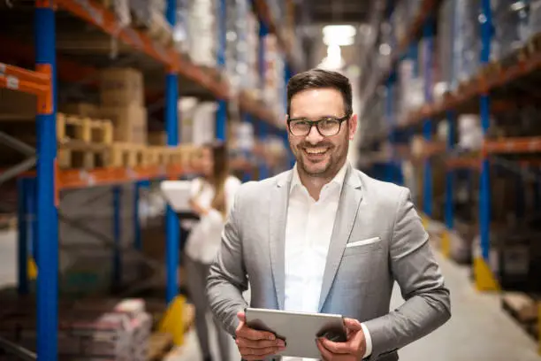 Photo of Portrait of successful middle aged Caucasian manager businessman holding tablet computer in large warehouse organizing distribution. Business people.
