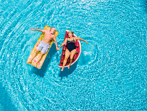 Above vertical view of people old senior couple taking hands with love and having fun on the blue clear swimming pool together enjoying the summer holiday vacation with trendy coloured lilos inflatable mattress