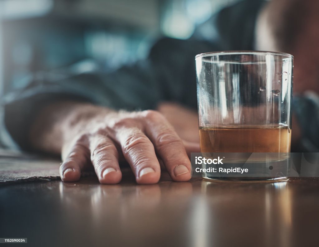 Hand by glass of liquor, man's head on table A man seems to have passed out or be sleeping, his head on a table, his hand near a glass of an alcoholic drink. Alcohol Abuse Stock Photo
