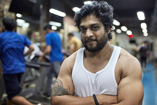 Portrait of confident muscular man with arms crossed at health club. Bearded male is in white sleeveless t-shirt. He is standing in gym.
