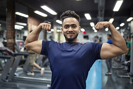 Portrait of young muscular man flexing muscles with pride at gym. Smiling macho male is standing at health club. He is in blue t-shirt.