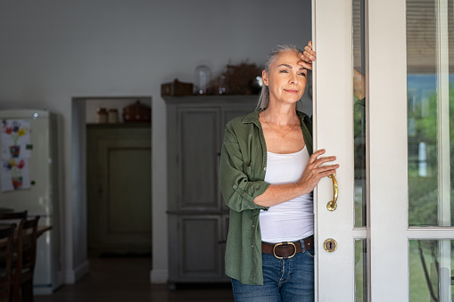 Thoughtful senior woman standing at home entrance leaning against door. Mature woman waiting husband at doorway and looking away. Lady in casual clothing standing at door and thinking.