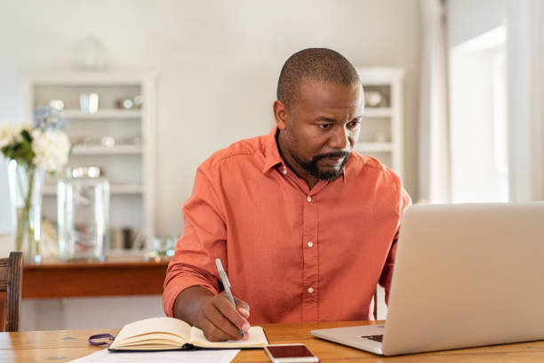 Mature man working on laptop at home Mature man working on laptop while taking notes. Businessman working at home with computer while writing on agenda. African man managing home finance, reviewing bank account and using laptop in living room. middle age adult studying stock pictures, royalty-free photos & images