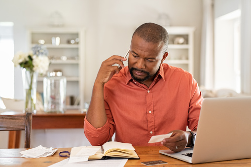 Mature man paying bills with laptop while talking on phone. Thoughtful man at home in conversation over smartphone while checking receipts. Worried african man discussing expenses over phone with bank insurance.
