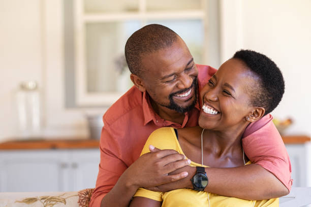 Mature black couple in love laughing Mature black couple embracing on sofa while looking to each other. Romantic black man embracing woman from behind while laughing together. Happy african wife and husband loving in perfect harmony. wife stock pictures, royalty-free photos & images