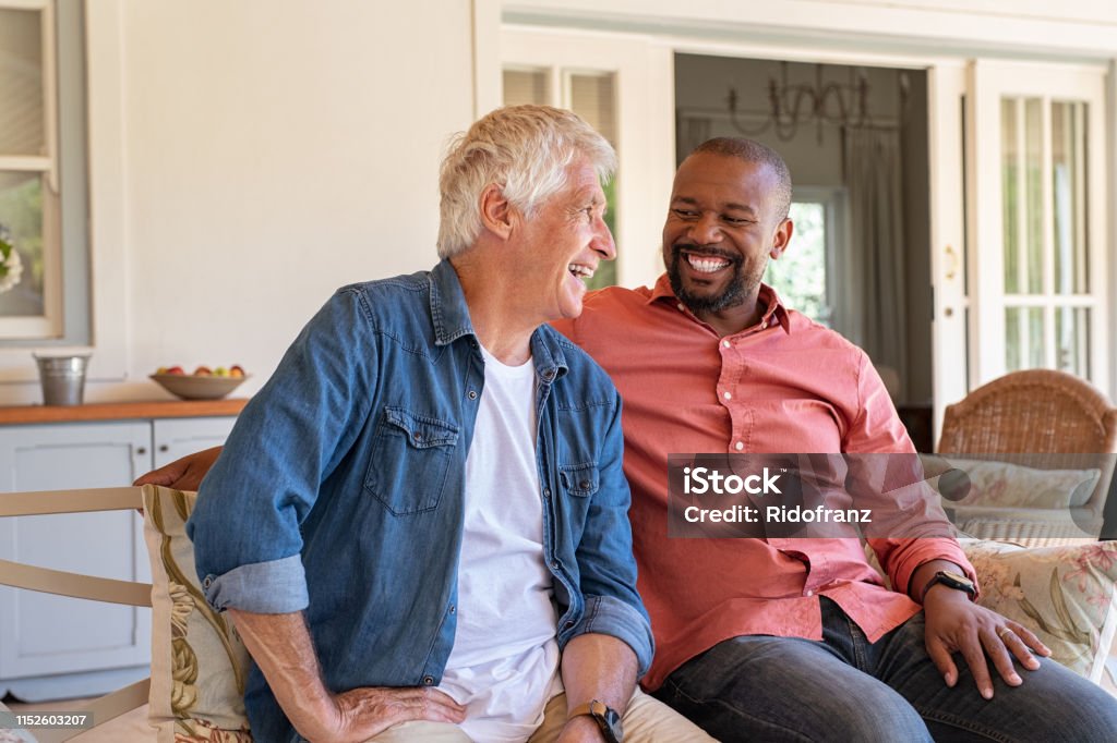 Happy men in a conversation Two friends talking while sitting on couch in the courtyard. Senior man and african guy laughing while in conversation sitting outside home. Two men talking to each other and enjoying. Friendship Stock Photo