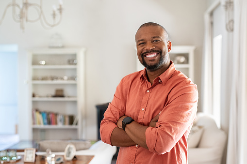 Portrait of smiling mature man with beard standing with crossed arms. Confident african man with folded arms at home looking at camera. Happy senior in casual feeling good.
