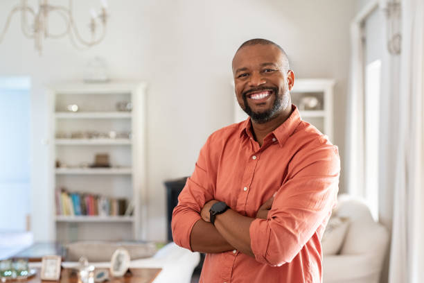 Mature black man with crossed arms looking at camera Portrait of smiling mature man with beard standing with crossed arms. Confident african man with folded arms at home looking at camera. Happy senior in casual feeling good. mature men stock pictures, royalty-free photos & images