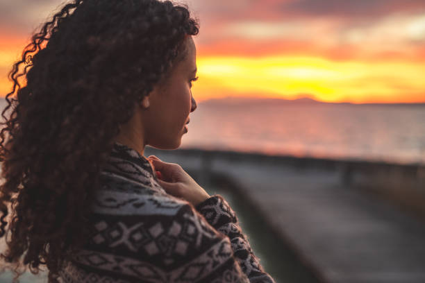 Thoughtful evening mood with a young afro latino woman Young African American woman is standing on the promenade at the lake, looking thoughtfully towards the water and the setting sun. The girl in a vest is in a thoughtful mood african sunset stock pictures, royalty-free photos & images