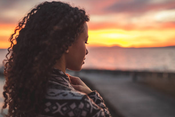 Thoughtful evening mood with a young afro latino woman Young African American woman is standing on the promenade at the lake, looking thoughtfully towards the water and the setting sun. The girl in a vest is in a thoughtful mood afro latinx ethnicity stock pictures, royalty-free photos & images