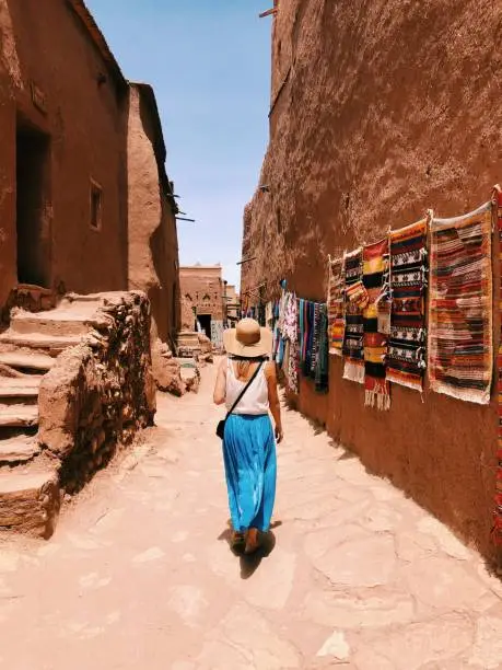 A young woman wearing a long blue skirt, white jersey and wicker hat is walking along a street in the Ait Ben Haddou village, hung with traditional Moroccan carpets. Blue cloudless sky on the background.