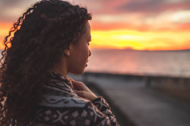 Thoughtful evening mood with a young afro latino woman Young African American woman is standing on the promenade at the lake, looking thoughtfully towards the water and the setting sun. The girl in a vest is in a thoughtful mood afro latinx ethnicity stock pictures, royalty-free photos & images