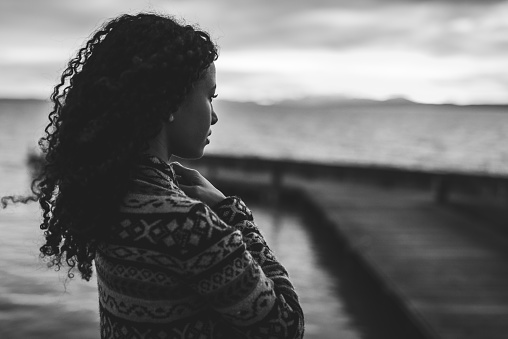 Young African American woman is standing on the promenade at the lake, looking thoughtfully towards the water and the setting sun. The photo in black and white