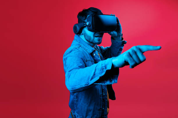 istock Young handsome bearded man wearing virtual reality goggles in studio on red background in neon light. Smartphone using with VR headset. 1152611741