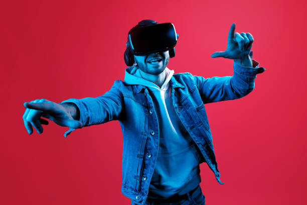 istock Young handsome bearded man wearing virtual reality goggles in studio on red background in neon light. Smartphone using with VR headset. 1152611764