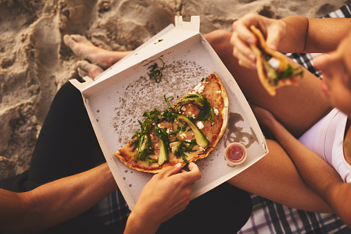 Shot of a young unrecognizable couple having a small picnic date and sharing a pizza on the beach at sunset