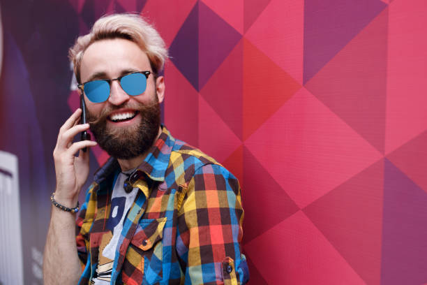 Bearded trendy stylish positive cheerful man wearing casual shirt, sun glasses, talking on mobile, over multicolore violet purple red background, empty blank copy space. stock photo