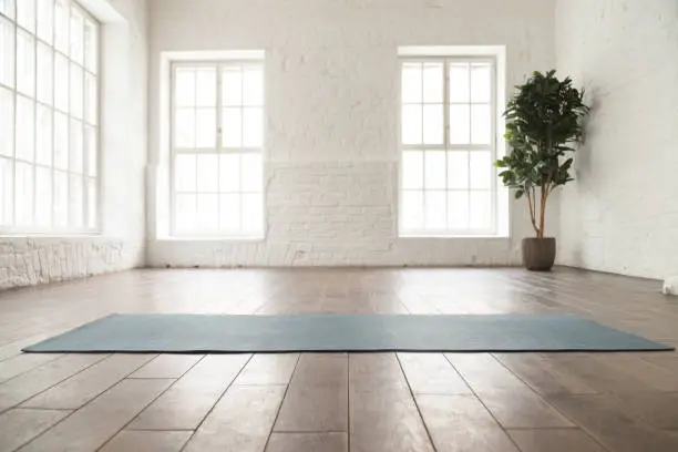Unrolled yoga mat on wooden floor in modern fitness center or at home with big windows and white brick walls, comfortable space for doing sport exercises, meditating, yoga equipment