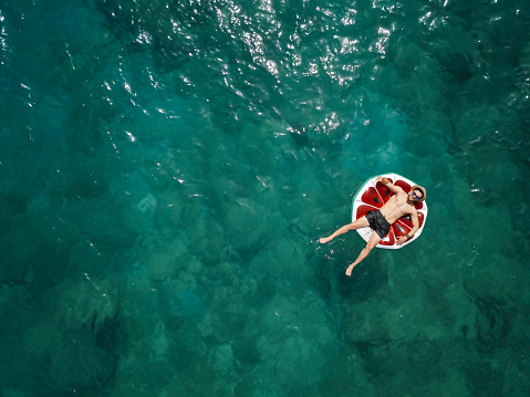 Young man with hat and sunglasses relaxing on watermelon shaped inflatable bed on the sea. Aerial drone footage. Turquoise mediterranean sea in Turkey, Alanya.