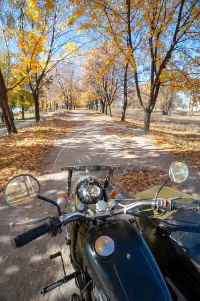 Vintage green motorcycle with a sidecar, view from the driver's seat through a transparent plexiglass flap on an autumn road. Chrome steering elements, fuel cap, front linkage shock absorbers.
