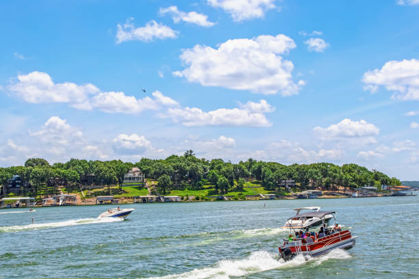 Pontoon boat full of people and two speedboats race down lake with luxury homes and docks on shore under bright blue sky with clouds and helicopter above stock photo