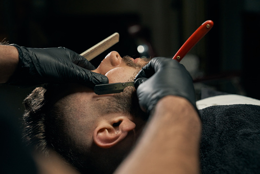 Close up of barbers hands in black gloves shaving bearded client with sharp red razor. Handsome bearded man is getting shaved by hairdresser at the barbershop.Concept of professional customer service