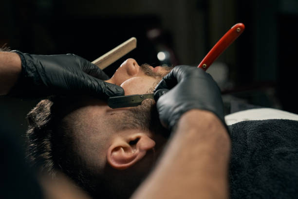 Handsome bearded man is getting shaved by hairdresser Close up of barbers hands in black gloves shaving bearded client with sharp red razor. Handsome bearded man is getting shaved by hairdresser at the barbershop.Concept of professional customer service razor blade photos stock pictures, royalty-free photos & images