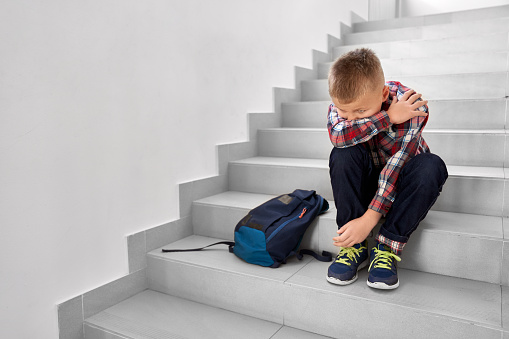 Little offended school boy sitting alone on stairs and hiding face from hands. Blond little boy sadly crying in school corridor, near backpack. Concept of bed marks.
