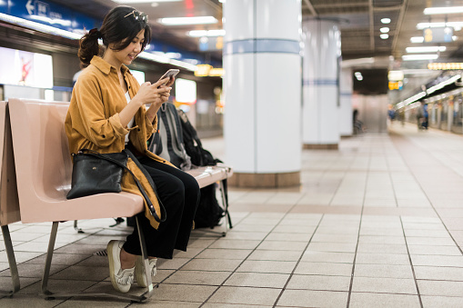 Young woman sitting on subway station bench texting friends while waiting for the train to arrive.Profile view looking down platform.