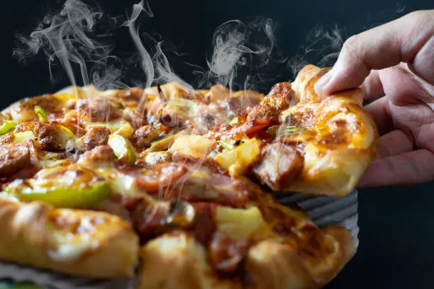 Photo of Hand picking sliced Pizza with cheese ham bacon and pepperoni on black background with hot steaming smoke. Food and cooking concept. Lunch time serve and hungry theme. Pizza delivery service to home.