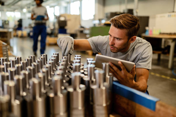 Young steel worker performing quality control of manufactured products in a warehouse. Young metal worker using touchpad while examining steel rods in a factory before the distribution. quality stock pictures, royalty-free photos & images