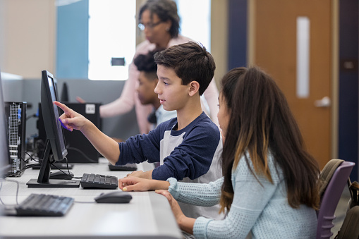 Mixed race male middle school student points to something on a computer monitor while working with his friend on an assignment. They are in the school's computer lab.