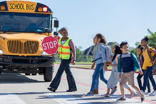 A group of school children cross the street with a senior female crossing guard.