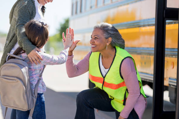 Senior female bus driver high-fives a new student A senior female bus driver kneels down to greet a new schoolgirl with a high-five. The new student is standing shyly beside her mother. custodian stock pictures, royalty-free photos & images