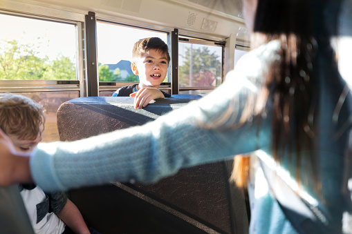 A schoolboy turns in his seat on the school bus and calls to his friend who is sitting across the bus from him.
