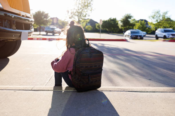 Anonymous schoolgirl sits on curb by herself An unknown schoolgirl with a backpack hugs her knees and sits on the curb by herself at a bus stop, while the bus is parked nearby. school exclusion stock pictures, royalty-free photos & images